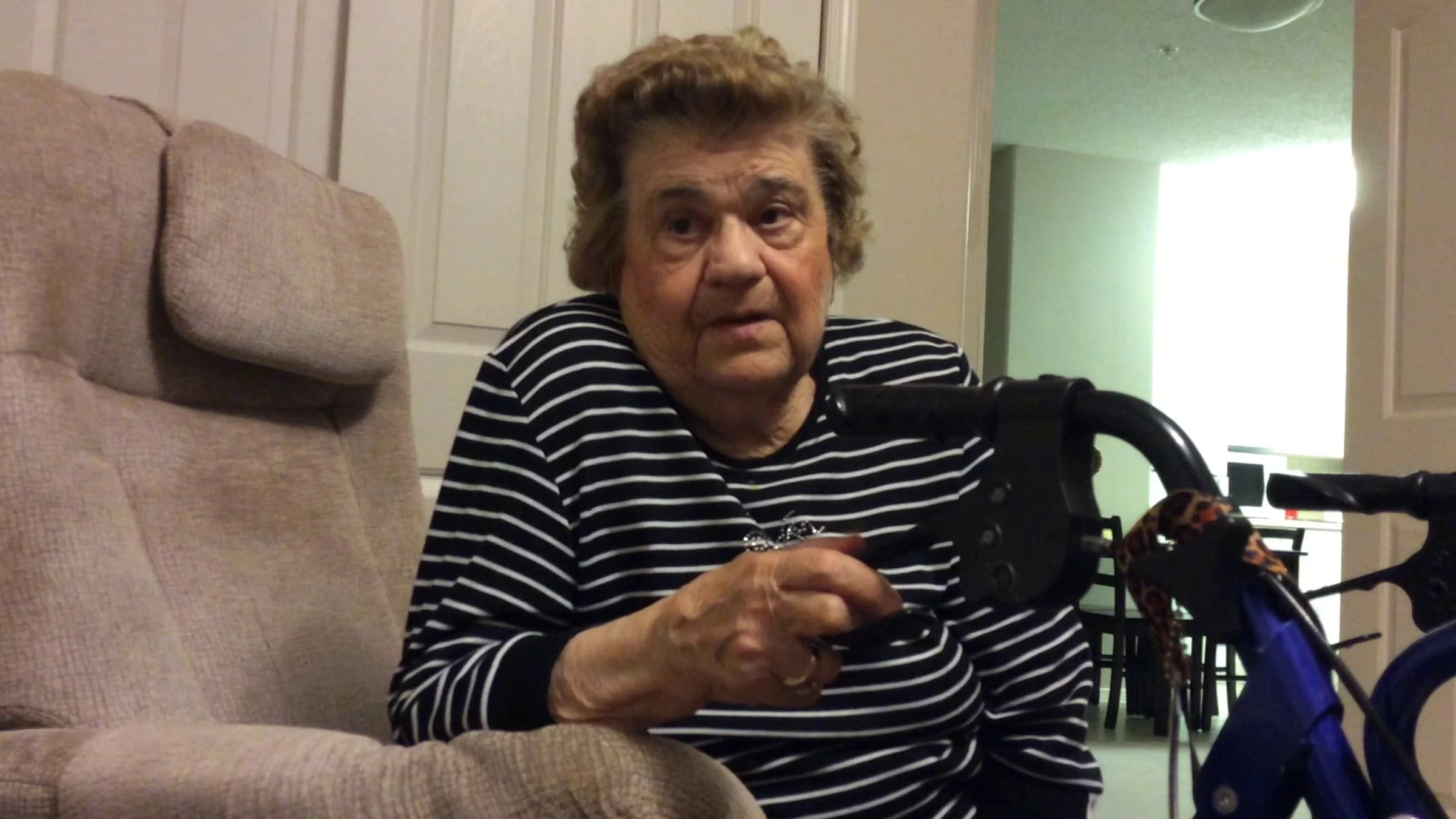 Elderly woman seated indoors in a wheelchair, wearing striped shirt, holding remote control, with chair and walker nearby in home setting.